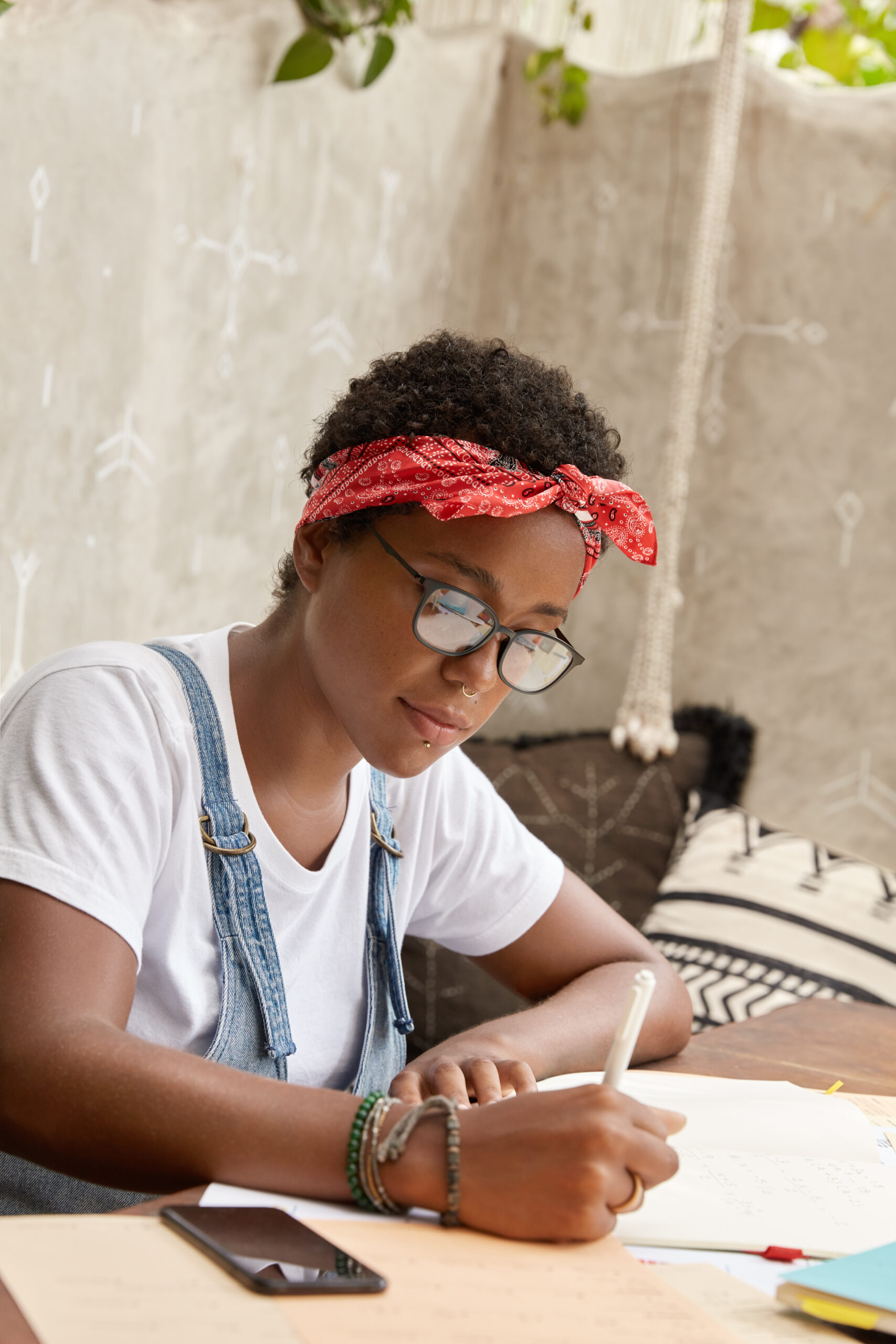 About busy student prepares for university seminar, writes down thesis, makes home assigment, prepares plan for presentation, studies marketing strategies, uses smart phone, wears piercing and bandana